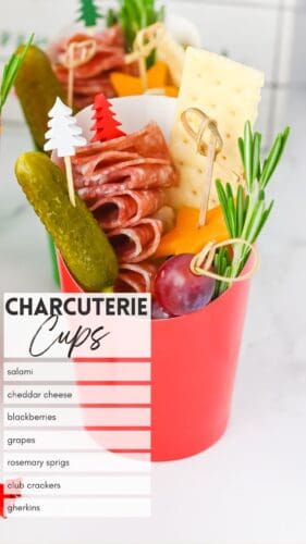 Christmas charcuterie cup containing salami, cheddar cheese, blackberries, grapes, crackers, rosemary, and a gherkin. Overlay text reads "Charcuterie Cups" with a labeled list: salami, cheddar cheese, blackberries, grapes, rosemary sprigs, club crackers, gherkins.