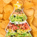 A close-up top view of a Christmas tree-shaped dish filled with layered taco dip, including refried beans, guacamole, sour cream, shredded cheese, diced tomatoes, olives, and green onions. A marbled cheese star tops the tree, and the dish is surrounded by round tortilla chips.