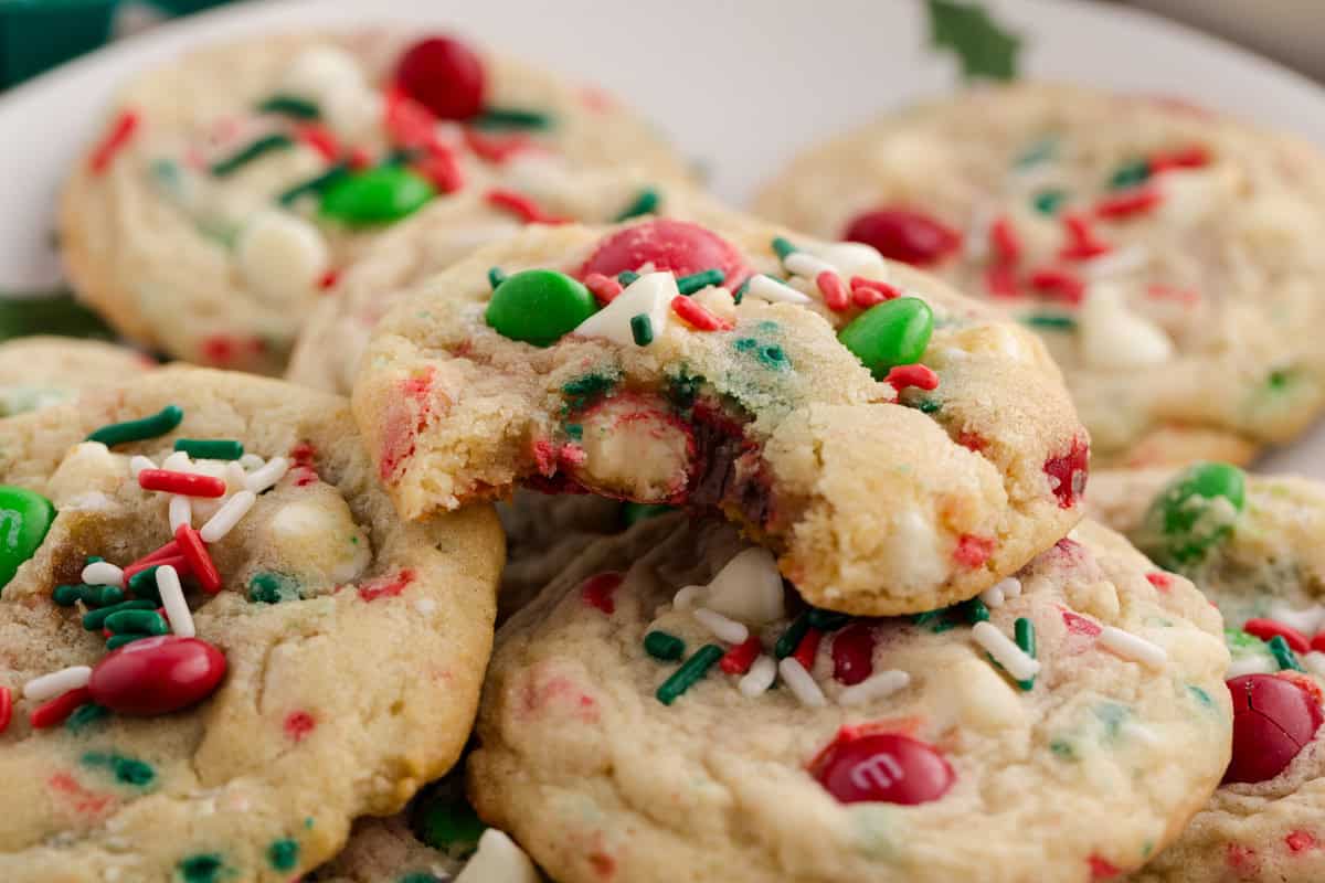Christmas cookies with red and green M&Ms, white chocolate chips, and festive sprinkles. One cookie has a bite taken out.