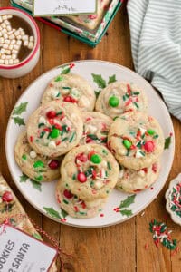 Overhead view of Christmas cookies with white chocolate chips, M&Ms, and sprinkles on a plate with holiday decor.