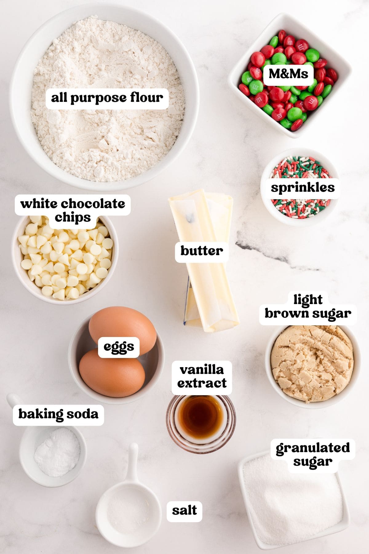 Top-down view of ingredients labeled with text: all-purpose flour, M&Ms, white chocolate chips, sprinkles, butter, light brown sugar, granulated sugar, eggs, vanilla extract, baking soda, and salt. Each ingredient is in its own bowl on a white surface.