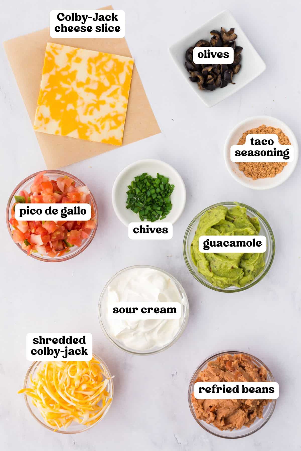 Overhead image of taco dip ingredients in small bowls, each labeled with text. Items include Colby-Jack cheese slice, olives, taco seasoning, chives, guacamole, sour cream, shredded Colby-Jack, pico de gallo, and refried beans.