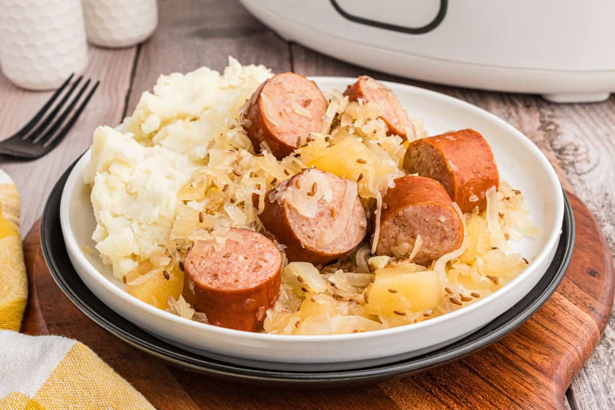 Sliced polish kielbasa and sauerkraut served with mashed potatoes. A slow cooker is n the background.