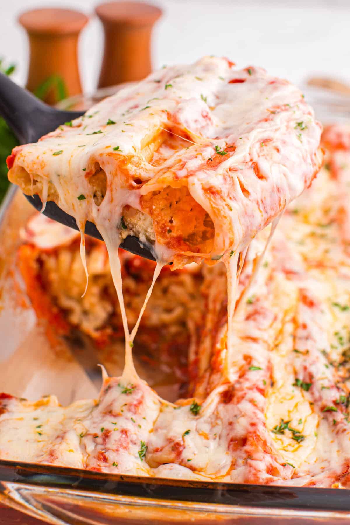 A close-up of a spatula lifting two stuffed manicotti from a baking dish, with stretchy melted cheese and a visible creamy filling inside the pasta. The dish is surrounded by more baked pasta and cheese.