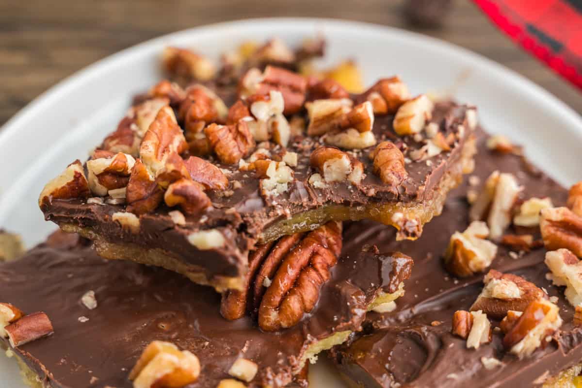 Close-up of saltine toffee on a white plate showing a piece with a bite taken out, revealing gooey toffee beneath the chocolate and pecan topping