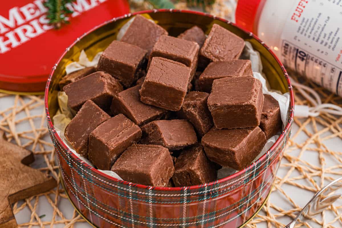 A round red plaid holiday tin filled to the top with square chocolate marshmallow fluff fudge pieces. A jar of Marshmallow Fluff and a red "Merry Christmas" lid in the background.