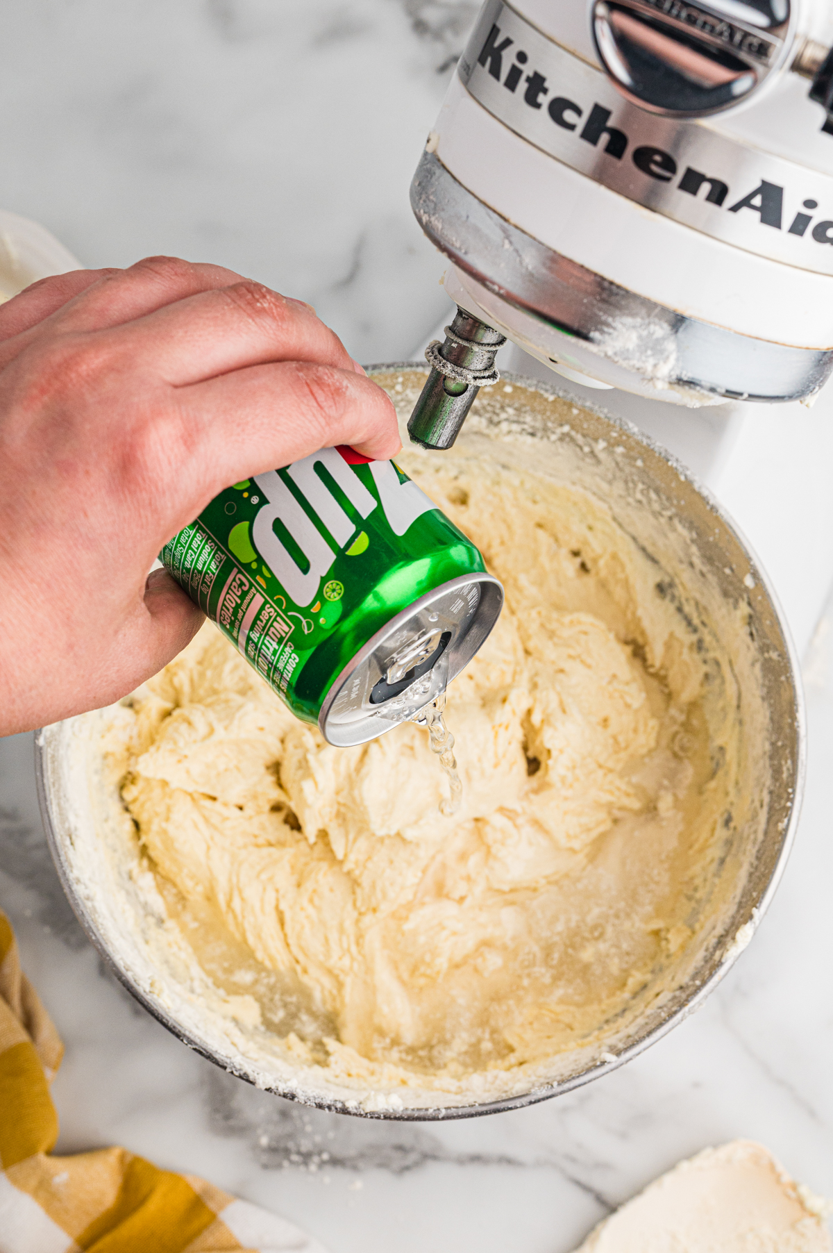 Hand pouring 7 Up into cake batter in mixer bowl.