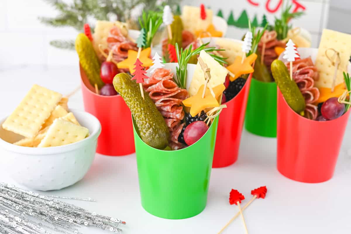 Red and green paper charcuterie cups are displayed together, each filled with folded salami skewers, cheddar cheese stars, grapes, gherkins, crackers, and rosemary.