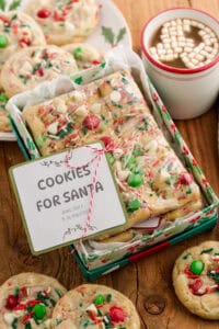 A holiday gift box filled with rectangular cookie bars topped with red and green M&Ms, white chocolate chips, and sprinkles, wrapped in plastic and tied with red and white string. A label reads “COOKIES FOR SANTA – BAKE 350 F, 11–14 MINUTES.” The box is surrounded by whole cookies, a holly-decorated plate, and a white mug of hot cocoa topped with mini marshmallows.
