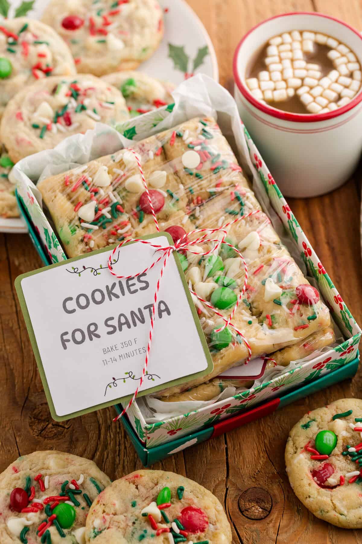 A holiday gift box filled with rectangular cookie bars topped with red and green M&Ms, white chocolate chips, and sprinkles, wrapped in plastic and tied with red and white string. A label reads “COOKIES FOR SANTA – BAKE 350 F, 11–14 MINUTES.” The box is surrounded by whole cookies, a holly-decorated plate, and a white mug of hot cocoa topped with mini marshmallows.
