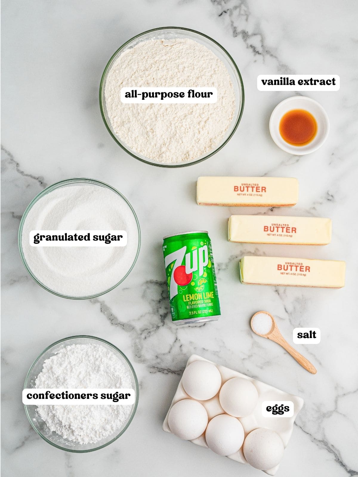 Labeled ingredients for 7 Up pound cake including: bowls of all-purpose flour, granulated sugar, confectioners sugar, and vanilla extract; a small wooden spoon of salt; six eggs in a carton; two sticks of unsalted butter; and a can of 7 Up.