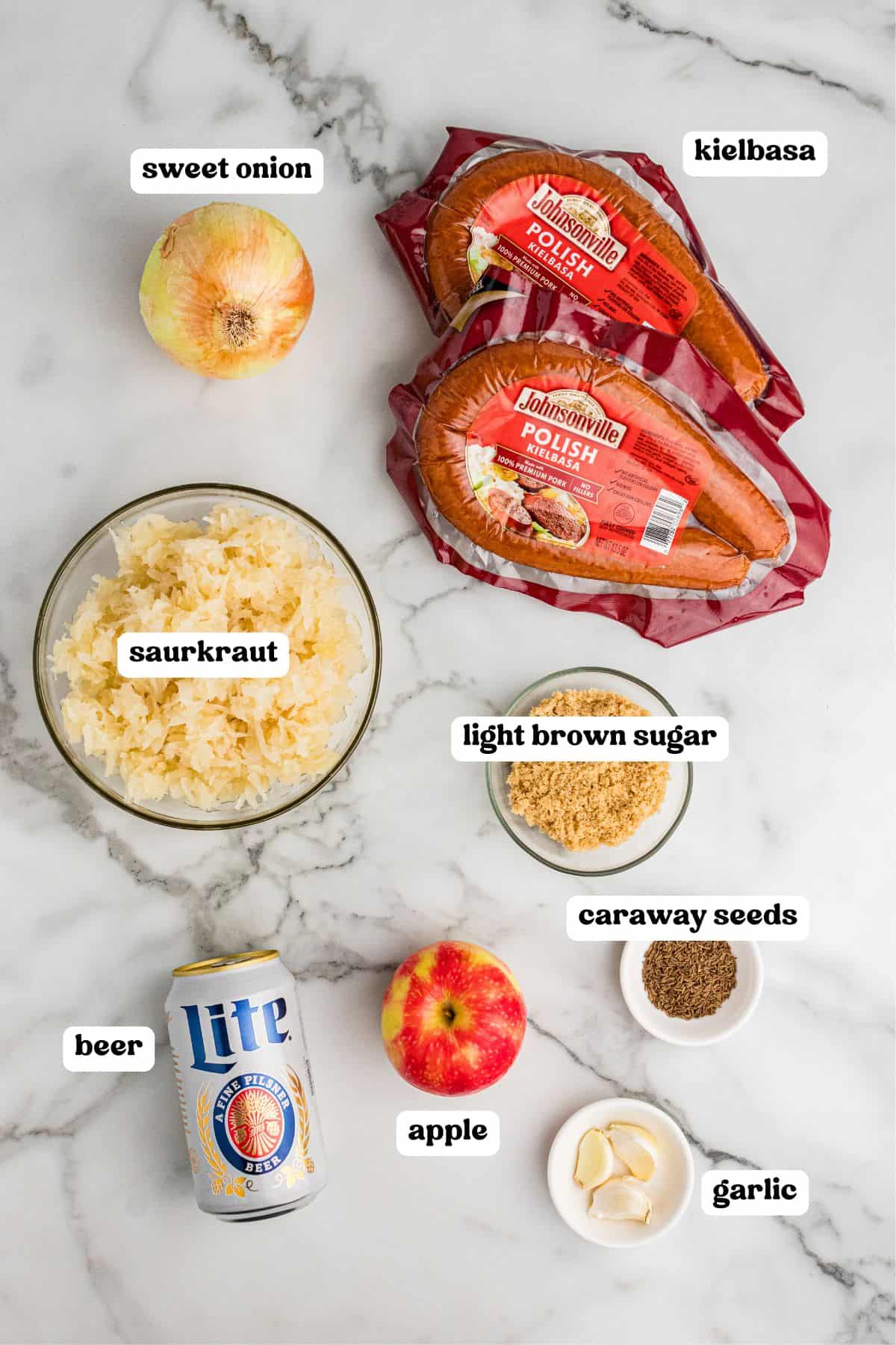 Ingredients on a white marble surface: sweet onion, sauerkraut, light brown sugar, caraway seeds, garlic cloves, an apple, a can of Lite beer, and two packages of Johnsonville Polish kielbasa.