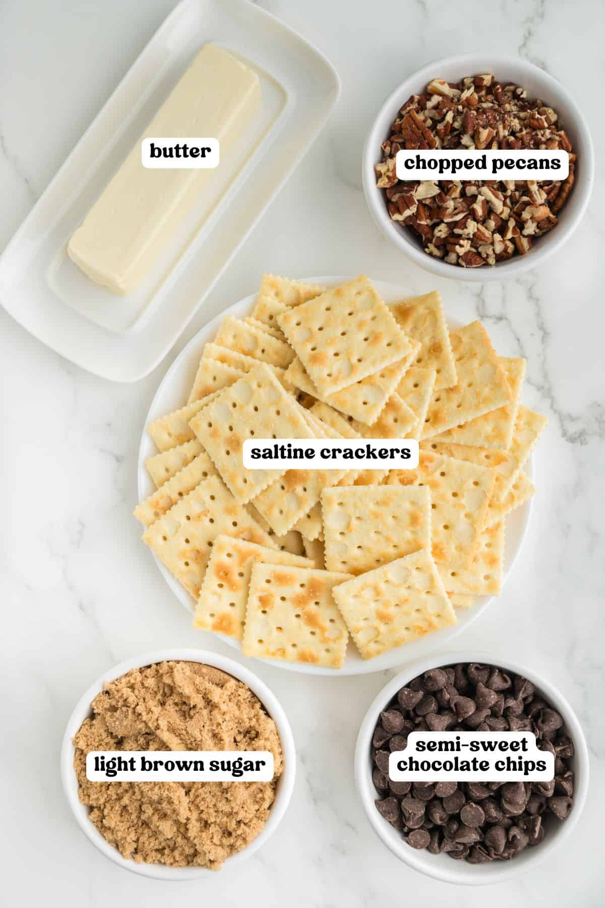 Top-down view of five labeled ingredients for saltine cracker toffee: a plate of saltine crackers, bowls of light brown sugar, semi-sweet chocolate chips, chopped pecans, and a stick of butter. 