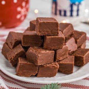 A stack of marshmallow fluff chocolate fudge pieces on a white plate placed on a red-striped cloth. In the background are festive decorations, including a red mug and a jar of Marshmallow Fluff.