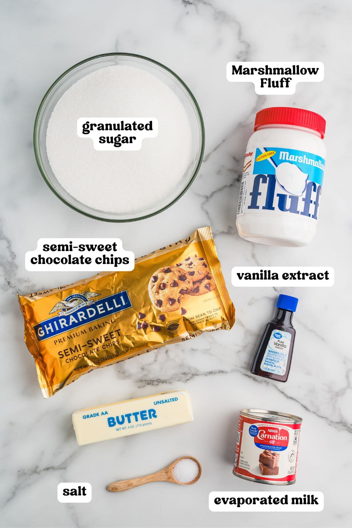 Overhead view of labeled ingredients for marshmallow fluff fudge on a marble surface. Items include a bowl of granulated sugar, a jar of Marshmallow Fluff, a bag of Ghirardelli semi-sweet chocolate chips, a bottle of vanilla extract, a stick of unsalted butter, a wooden spoon of salt, and a can of Carnation evaporated milk.