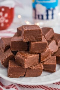 A stack of marshmallow fluff chocolate fudge pieces on a white plate placed on a red-striped cloth. In the background are festive decorations, including a red mug and a jar of Marshmallow Fluff.