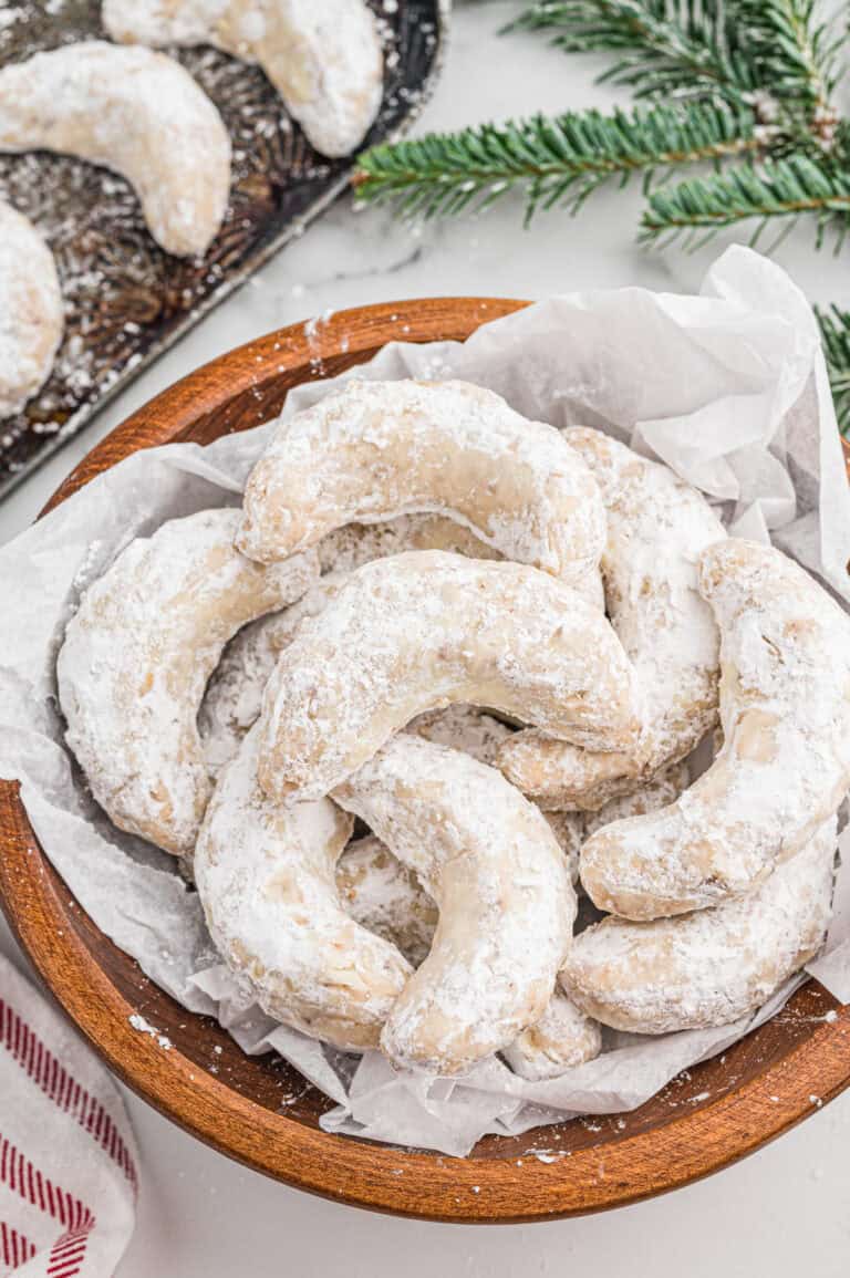 Top-down view of crescent-shaped cookies coated in powdered sugar, piled in a wooden bowl lined with white parchment paper.