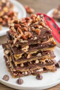 A stack of chocolate and pecan-topped saltine cracker toffee pieces on a white plate. Chocolate chips are scattered around the plate.