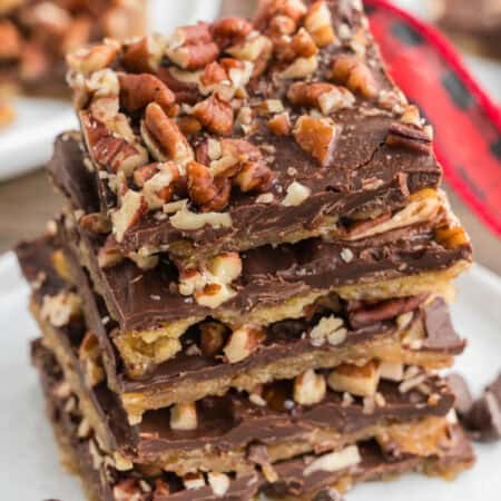A stack of chocolate and pecan-topped saltine cracker toffee pieces on a white plate. Chocolate chips are scattered around the plate.