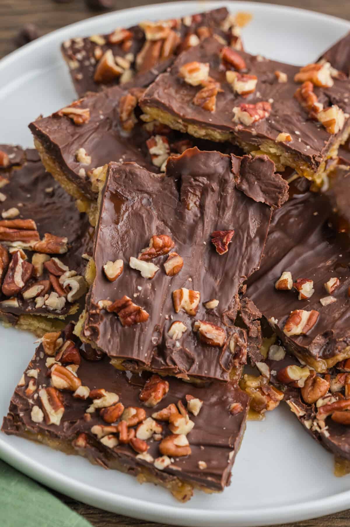A white plate filled with overlapping pieces of saltine cracker toffee. The toffee is coated in chocolate and topped with scattered chopped pecans.