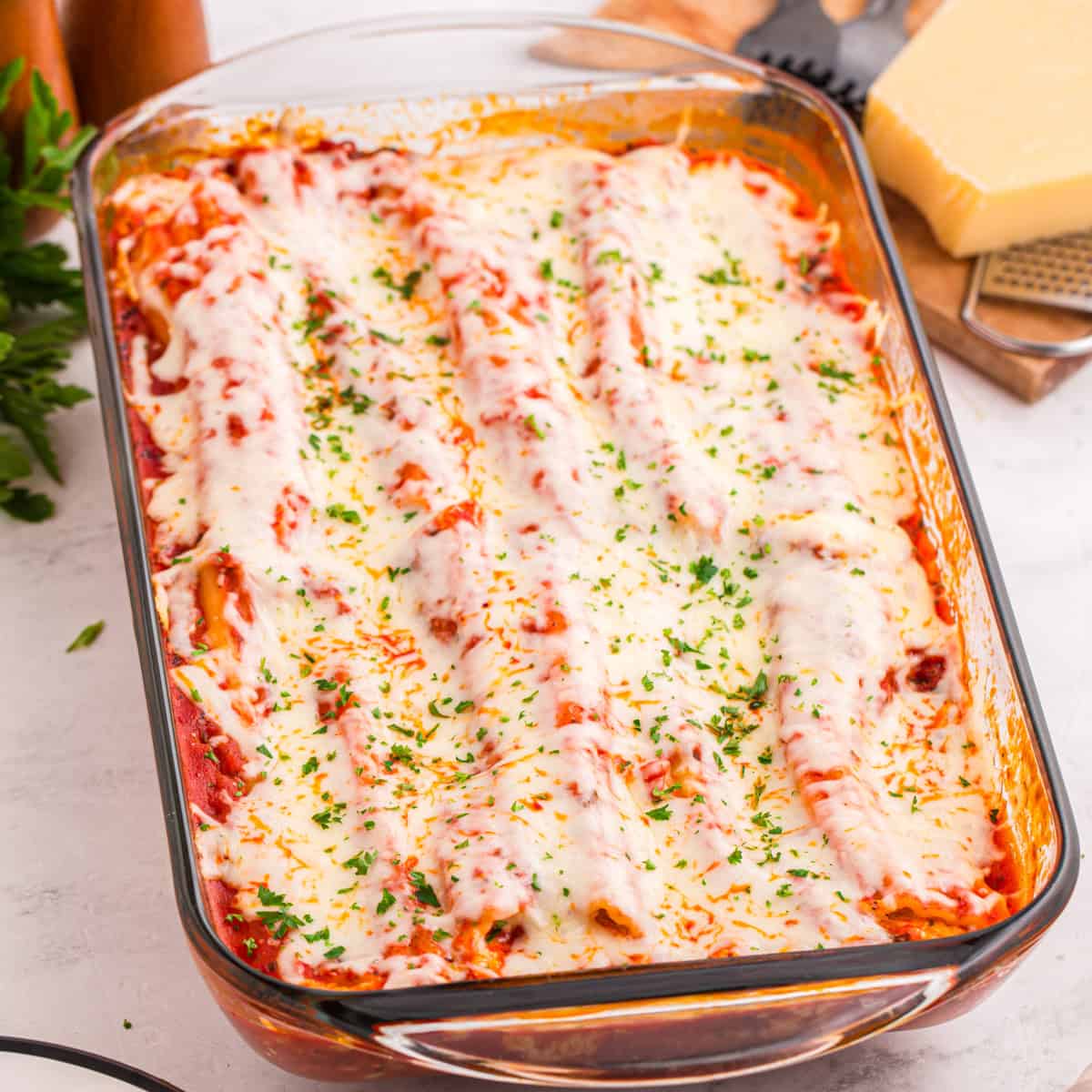 Baked manicotti covered in tomato sauce, melted mozzarella cheese, and garnished with chopped parsley in a glass baking dish. The cheese is slightly browned and bubbly on top.