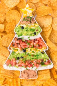 Overhead view of a full Christmas tree-shaped serving dish containing layered taco dip with refried beans, guacamole, sour cream, shredded cheese, diced tomatoes, olives, and green onions. A cheese star decorates the top, with tortilla chips surrounding the dish.