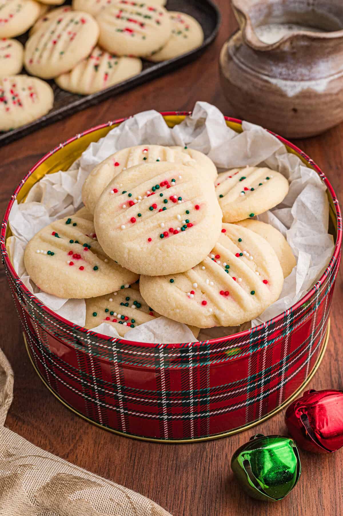 Round whipped shortbread cookies topped with red, green, and white sprinkles are arranged in a red plaid tin lined with white parchment paper. A tray of more cookies are in the background.