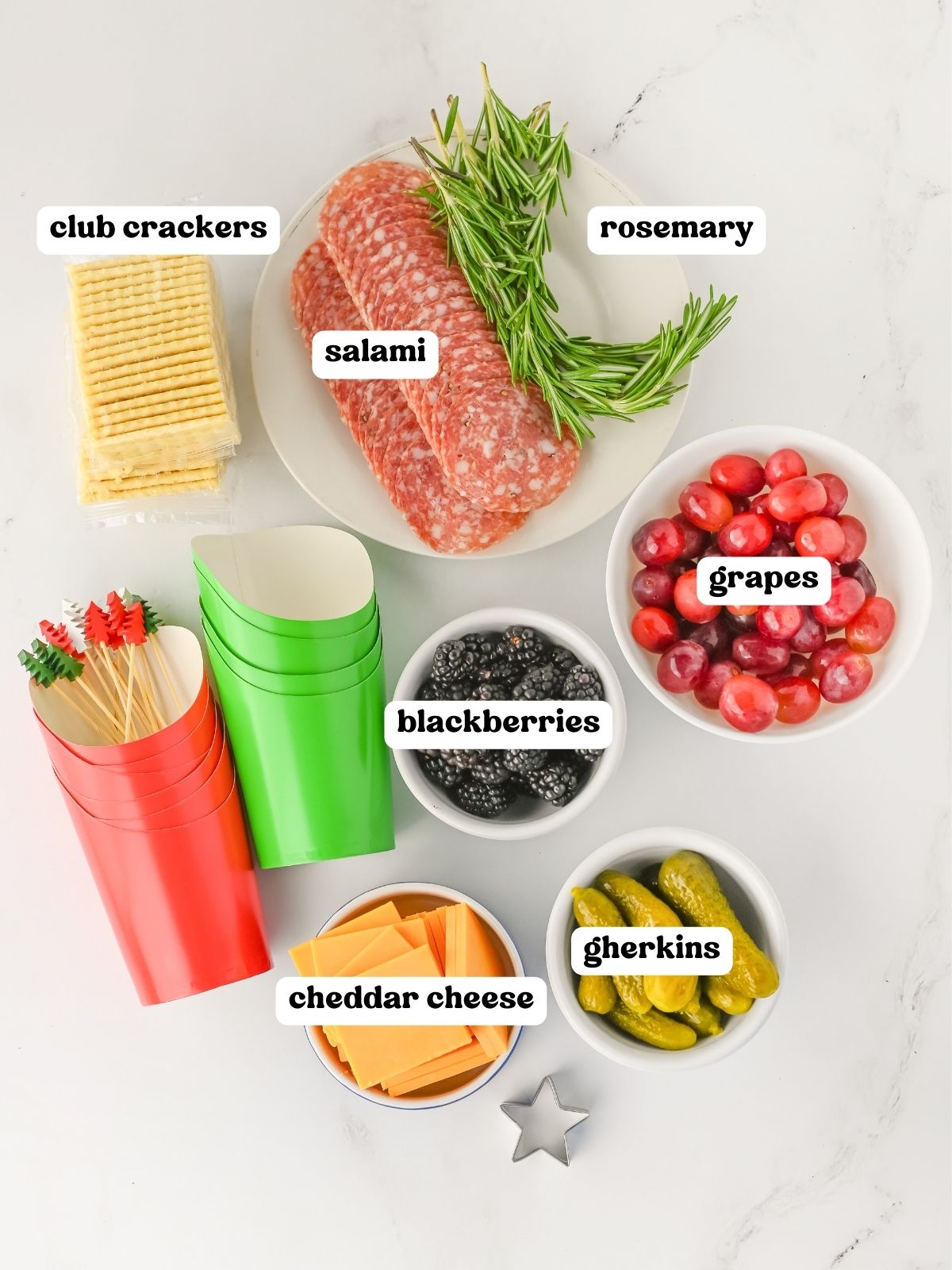 : Flat lay image showing labeled ingredients for charcuterie cups. Items include club crackers, rosemary sprigs, sliced salami on a plate, red grapes in a bowl, blackberries in a bowl, cheddar cheese slices in a bowl, and gherkins in a bowl. Red and green paper cups and decorative skewers are also visible.