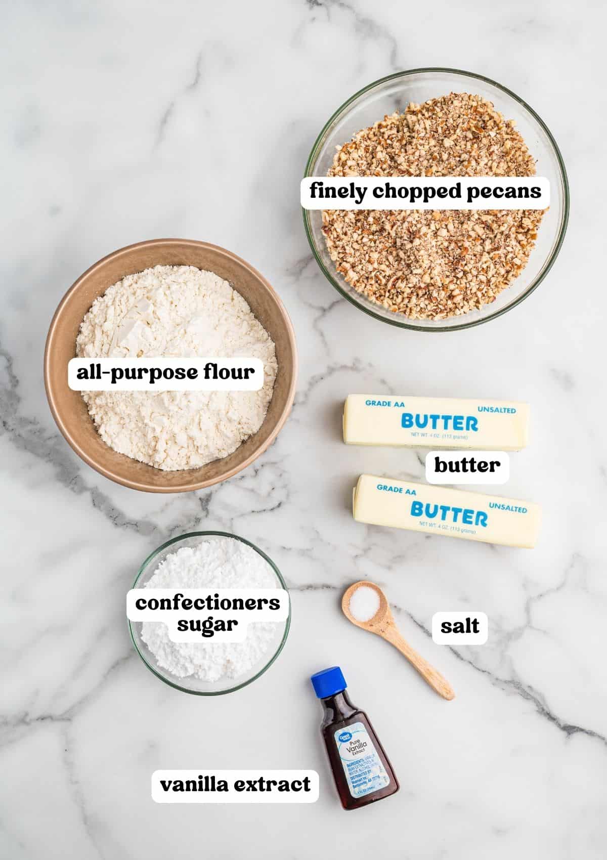 Overhead view of labeled ingredients for pecan crescent cookies, including bowls of all-purpose flour, finely chopped pecans, confectioners sugar, two sticks of unsalted butter, a small wooden spoon of salt, and a bottle of vanilla extract.