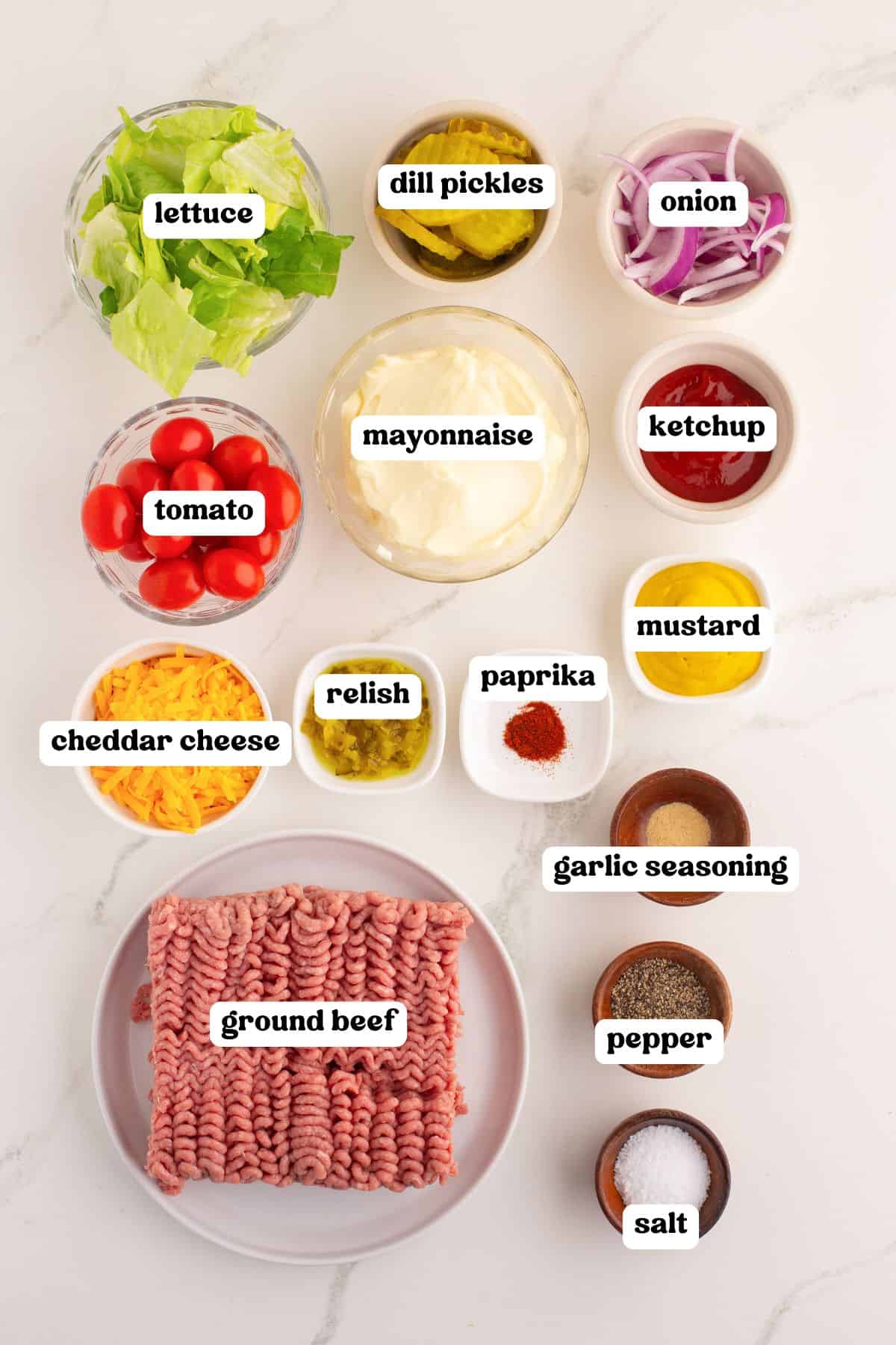 Flat lay of labeled ingredients for burger bowls including: lettuce, dill pickles, onion, tomato, mayonnaise, ketchup, mustard, relish, paprika, garlic seasoning, salt, pepper, cheddar cheese, and raw ground beef.