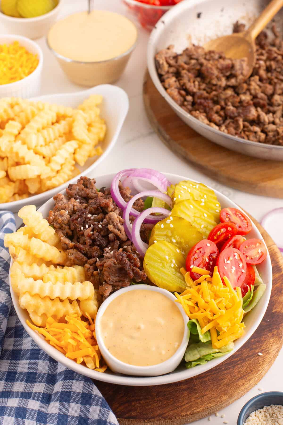 A burger bowl with crinkle-cut French fries, seasoned ground beef, sliced red onions, dill pickles, halved cherry tomatoes, shredded cheddar cheese, lettuce, and a side of creamy special sauce. The bowl is surrounded by dishes of cooked beef, shredded cheese, pickles, tomatoes, and extra fries.