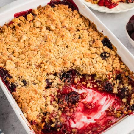 Baked cherry cobbler in a white rectangular dish, showing a golden crumbly topping and a missing portion revealing cherry filling.