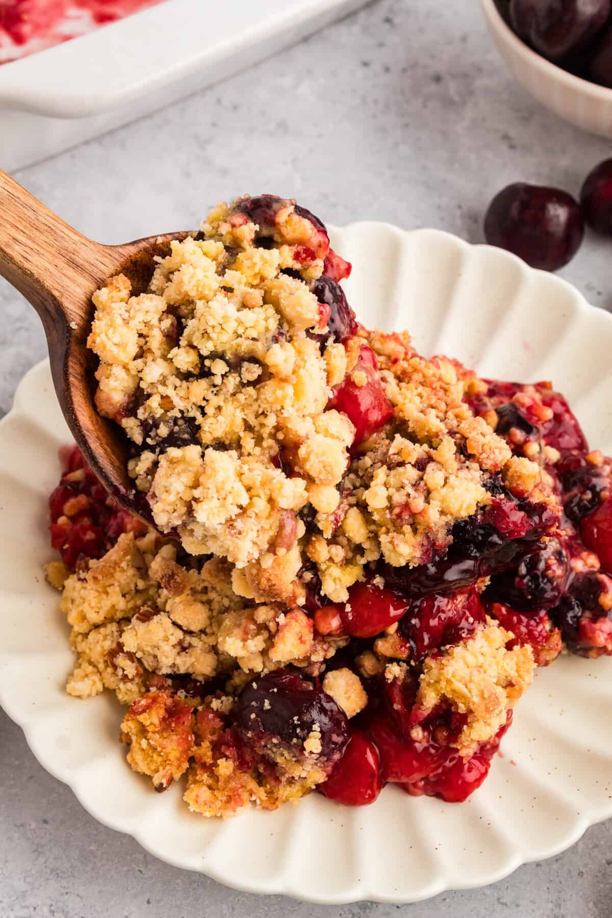 A wooden spoon scooping cherry cobbler onto a white scalloped plate. The cobbler has a crumbly topping with chunks of yellow cake mix and cherries visible in the filling.