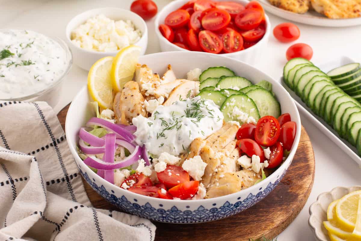 Chicken gyro bowl with grilled chicken slices, halved cherry tomatoes, cucumber slices, red onion, feta, and tzatziki sauce topped with dill. The bowl sits on a wooden board surrounded by small bowls of ingredients and a beige towel with black stripes.