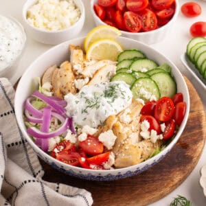 Chicken gyro bowl with grilled chicken, sliced cucumbers, cherry tomatoes, red onions, tzatziki sauce with dill, crumbled feta, and lemon slices.