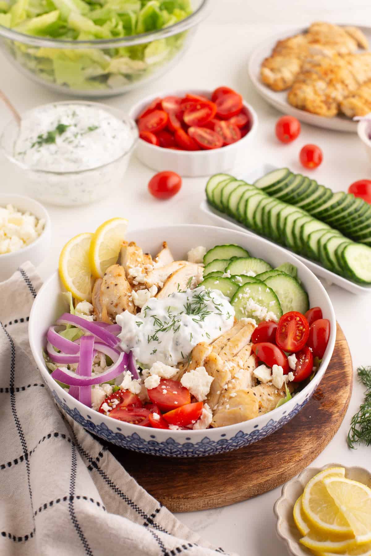 Chicken gyro bowl with grilled chicken, sliced cucumbers, cherry tomatoes, red onions, tzatziki sauce with dill, crumbled feta, and lemon slices. The background includes bowls of lettuce, cherry tomatoes, cucumber slices, feta, and cooked chicken.