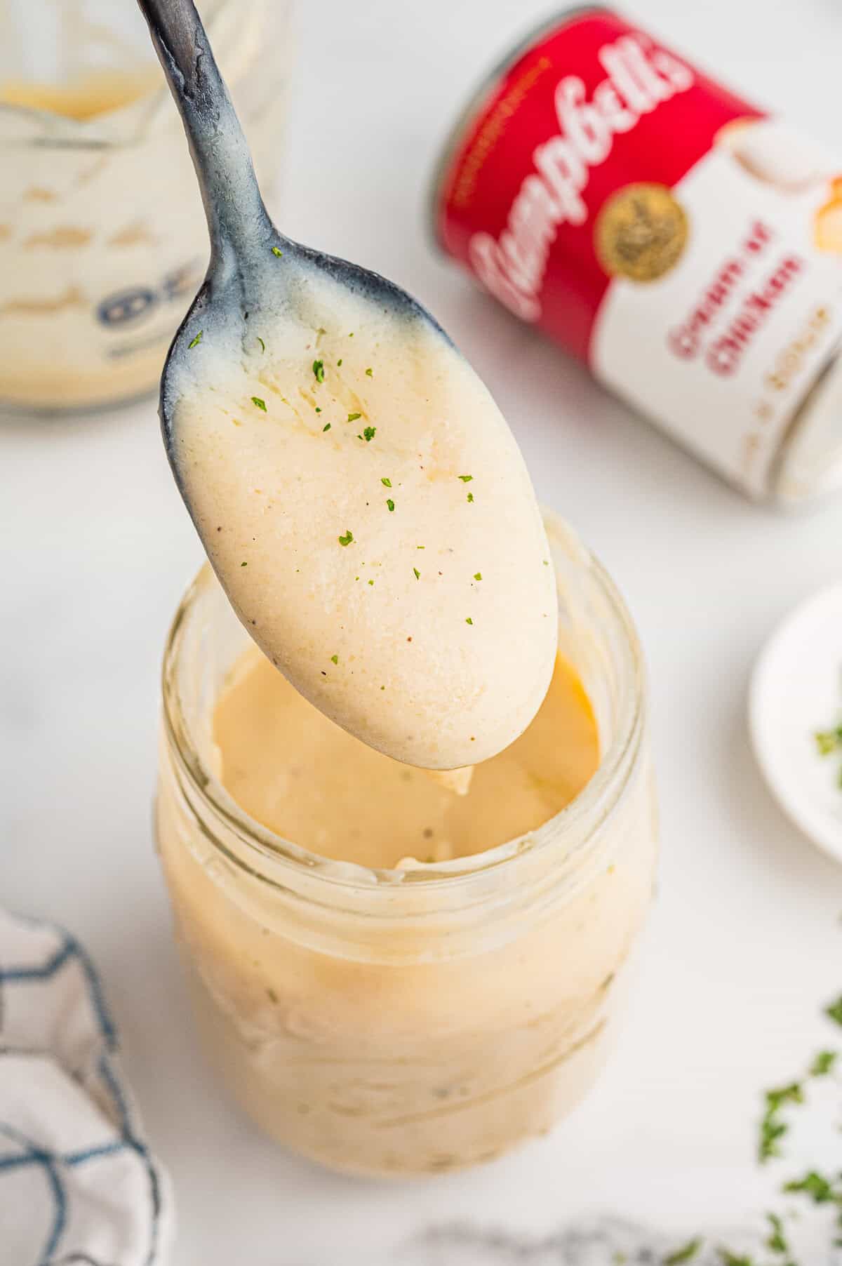 A spoon of homemade condensed cream of chicken soup above an open mason jar. The soup is lightly speckled with herbs. A Campbell&rsquo;s can and plate of parsley are visible in the background.
