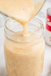 Cream of chicken soup being poured into a mason jar with a can of condensed cream of chicken soup in the background.