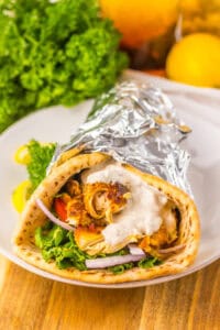 A slow cooker chicken shawarma wrap on a white plate, filled with greens, tomato, red onion, shredded chicken, and sauce, wrapped in foil. Garnished with parsley and lemon slices.