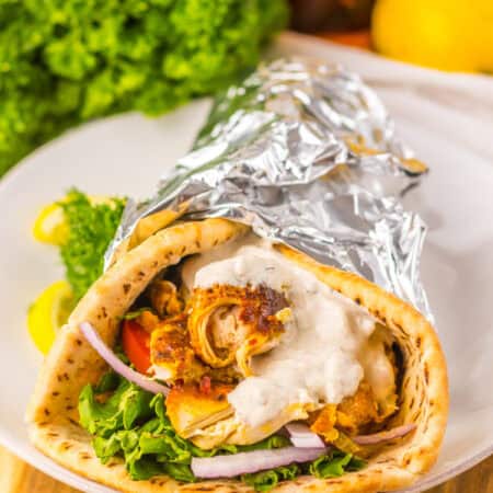 A slow cooker chicken shawarma wrap on a white plate, filled with greens, tomato, red onion, shredded chicken, and sauce, wrapped in foil. Garnished with parsley and lemon slices.