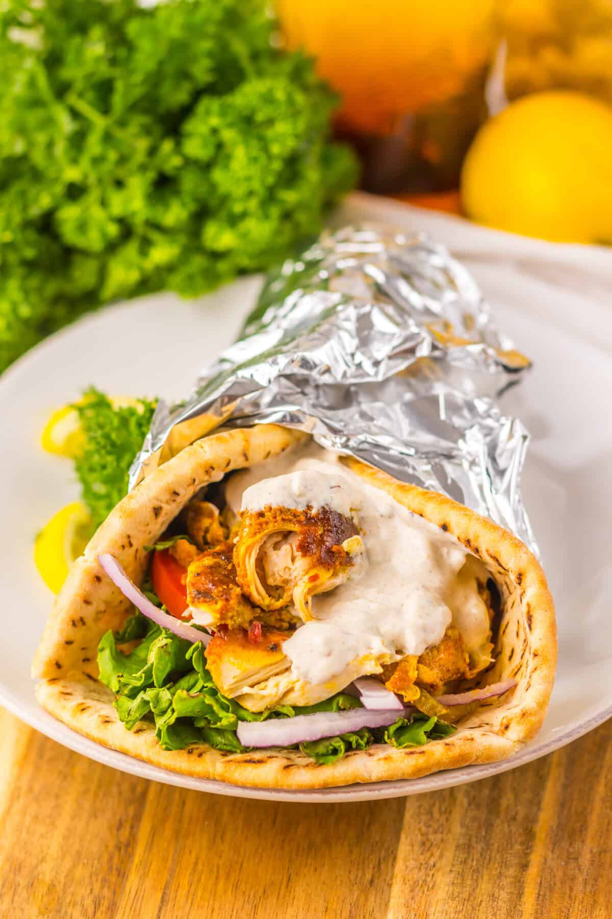 A slow cooker chicken shawarma wrap on a white plate, filled with greens, tomato, red onion, shredded chicken, and sauce, wrapped in foil. Garnished with parsley and lemon slices.