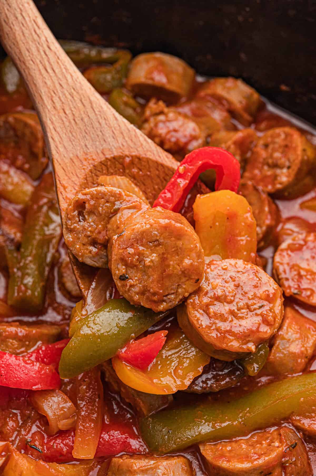 A wooden spoon holds sliced sausage, red and yellow bell pepper strips, and onion over a crockpot of sausage and peppers in marinara sauce.