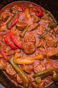 A close-up view of a cooked sausage and peppers dish in a slow cooker, showing sliced sausage rounds, red, yellow, and green bell pepper strips, and onions coated in thick marinara sauce.