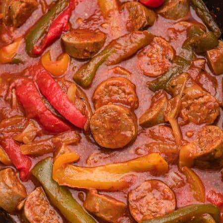 A close-up view of a cooked sausage and peppers dish in a slow cooker, showing sliced sausage rounds, red, yellow, and green bell pepper strips, and onions coated in thick marinara sauce.