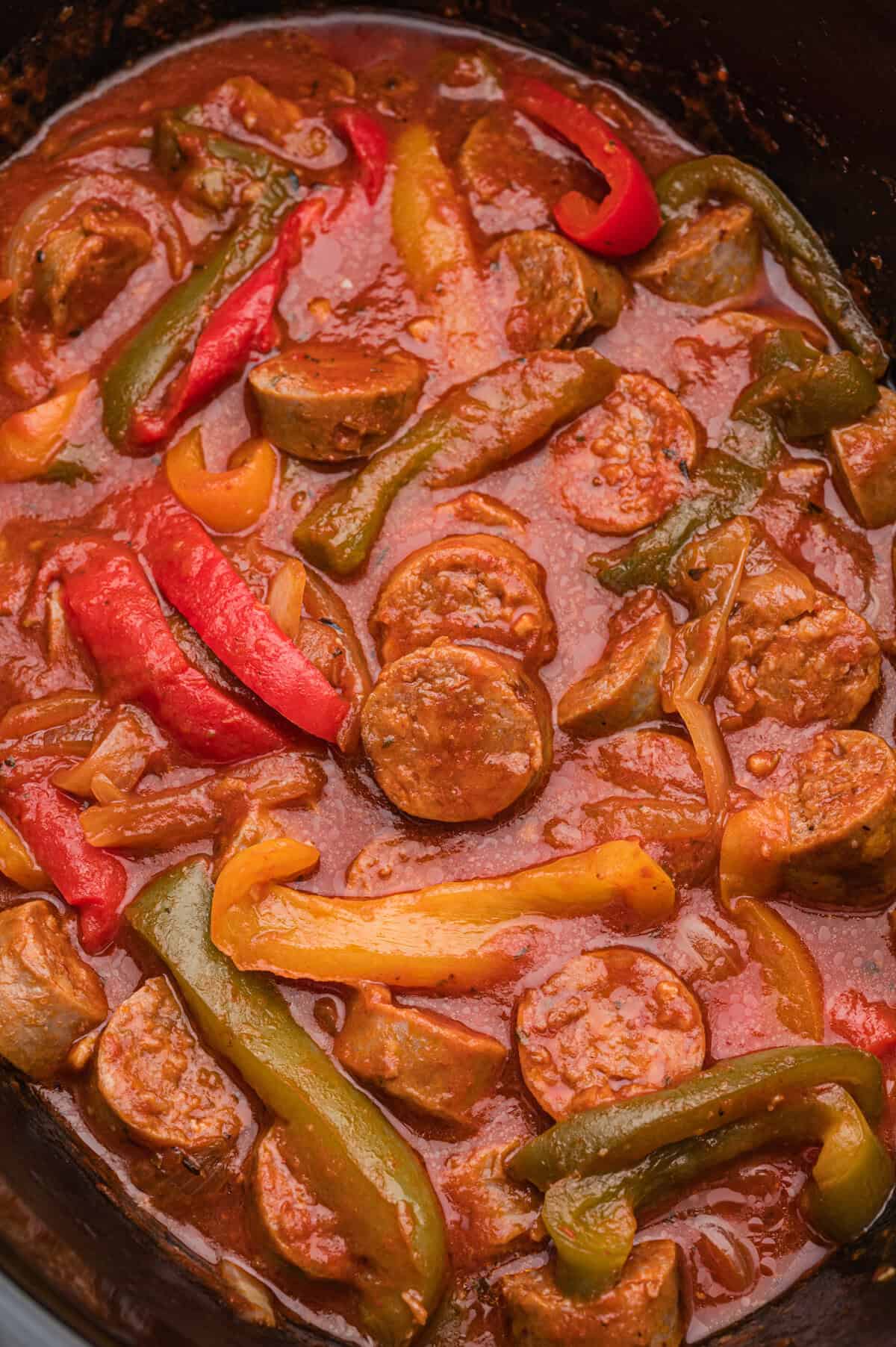 A close-up view of a cooked sausage and peppers dish in a slow cooker, showing sliced sausage rounds, red, yellow, and green bell pepper strips, and onions coated in thick marinara sauce.