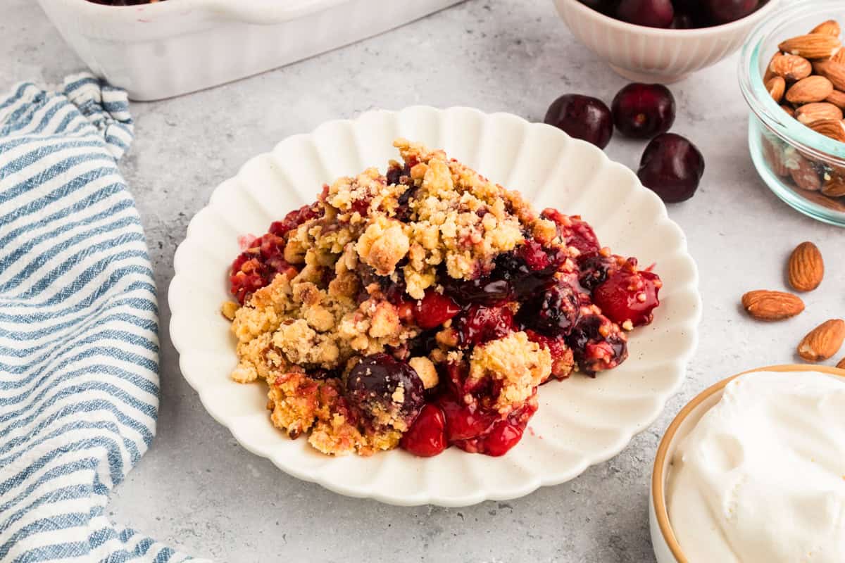 A serving of cherry cobbler from cake mix on a scalloped white plate, featuring a golden crumb topping and visible cherries