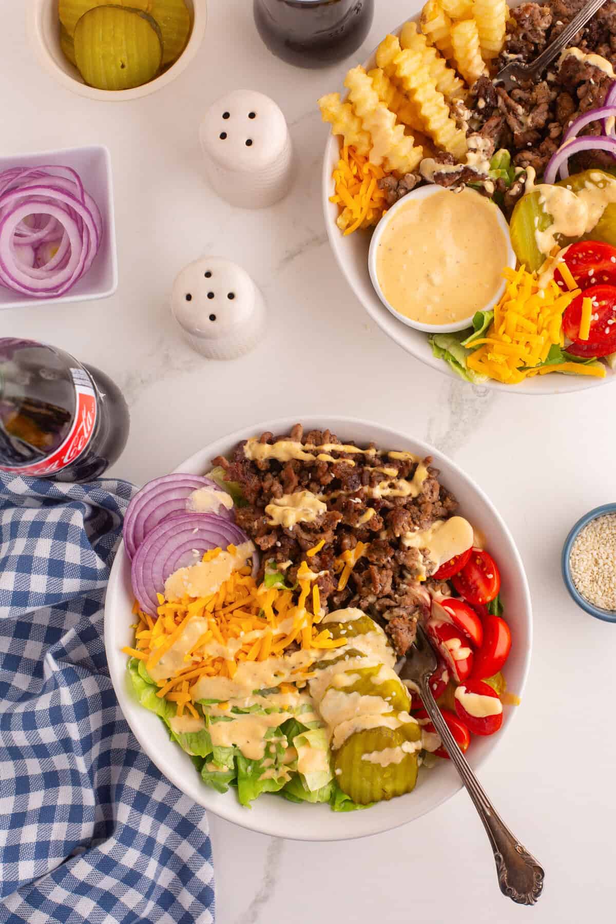 Overhead view of two burger bowls filled with cooked ground beef, shredded cheddar cheese, cherry tomatoes, crinkle-cut pickles, sliced red onion, chopped lettuce, and drizzled creamy dressing. Surrounding the bowls are a bottle of Coca-Cola, containers of pickles and onions, salt and pepper shakers, and a blue gingham napkin.