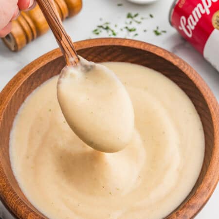 A hand holds a spoon scooping thick homemade cream of chicken soup from a wooden bowl. A red and white can of Campbell&rsquo;s cream of chicken soup ia in the background.