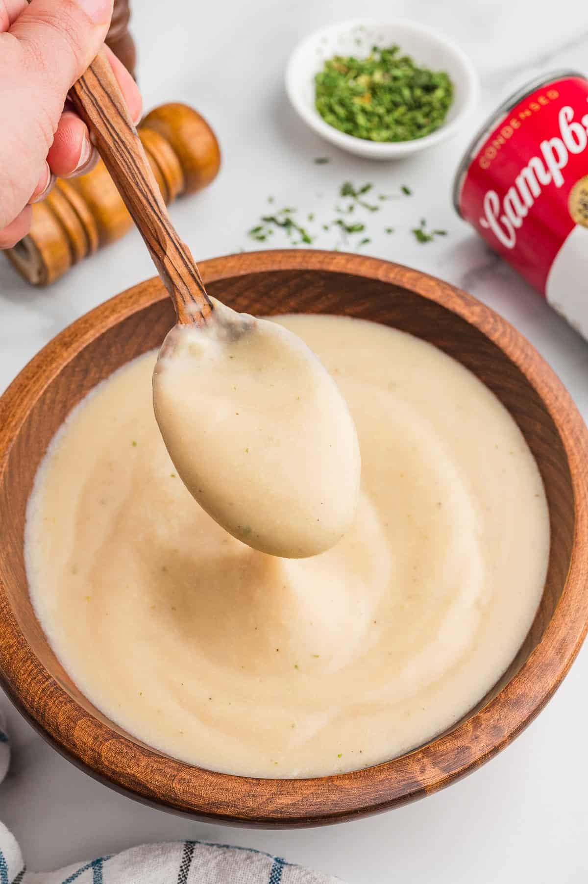 A hand holds a spoon scooping thick homemade cream of chicken soup from a wooden bowl. A red and white can of Campbell&rsquo;s cream of chicken soup ia in the background.