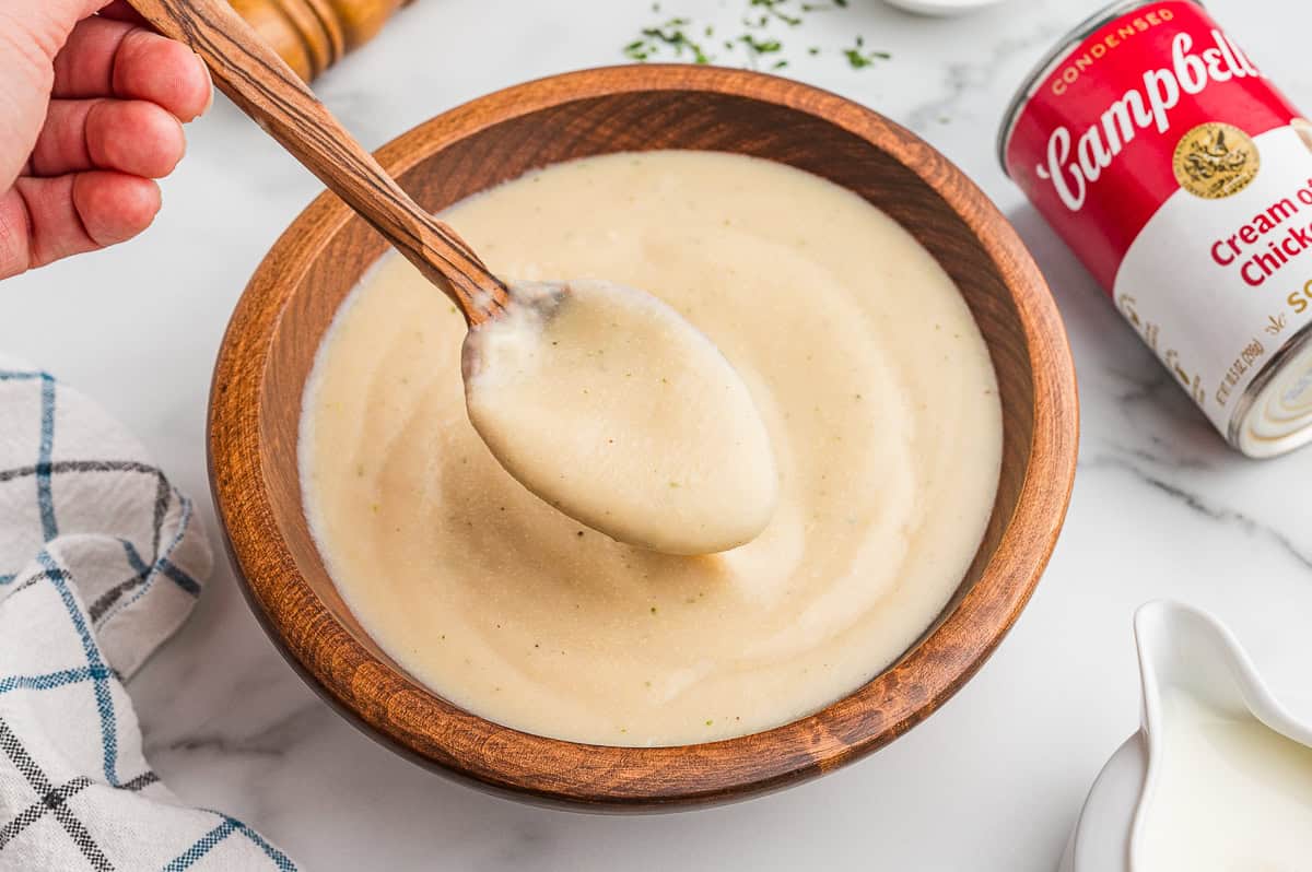 Bowl of cream of chicken soup with a wooden spoon resting in it. A Campbell&rsquo;s condensed cream of chicken soup can, chopped parsley, and kitchen towel surround the bowl.